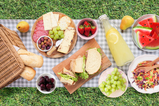 Summer picnic food scene. Top view with blanket over a green lawn background. Picnic basket, fruit, salad, sandwiches and snacks.