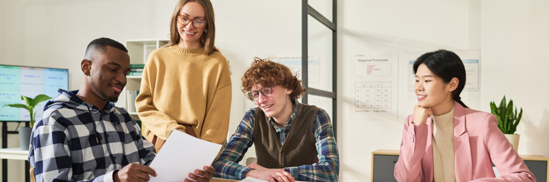 Group of students from diverse backgrounds studying foreign languages in classroom, collaborating on project. Teacher guiding students while reviewing documents, with charts and plants in background