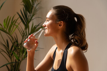 Fit woman in sportswear smiling and drinking water from a glass bottle after a workout. Natural light, tropical plant background. Healthy lifestyle and hydration concept.