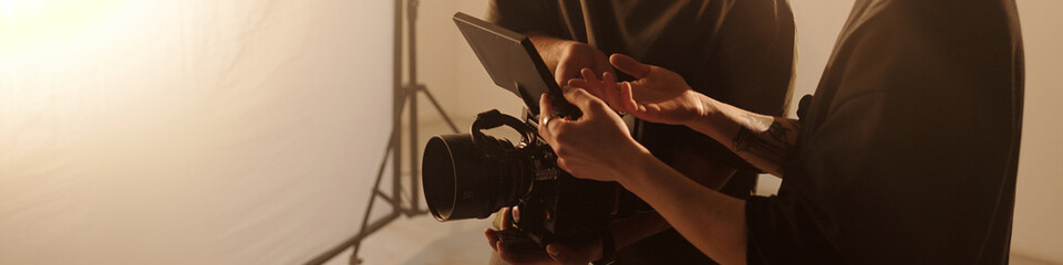 Header image of young cinematographer and assistant discussing camera settings while checking footage on monitor in studio