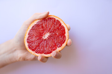 Close-up of a hand gently holding a juicy red grapefruit half on soft lilac background, symbolizing...