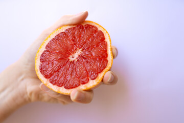 Close-up of a hand gently holding a juicy red grapefruit half on soft lilac background, symbolizing...