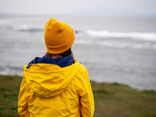 A woman in a yellow jacket stands on a grassy hill overlooking the ocean. The scene is peaceful and...