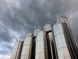Row of tall silver tanks are lined up in front of a cloudy sky
