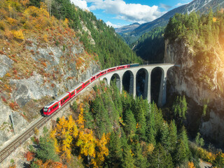 Aerial view of modern red train on Landwasser viaduct in alpine mountains, colorful forest at...