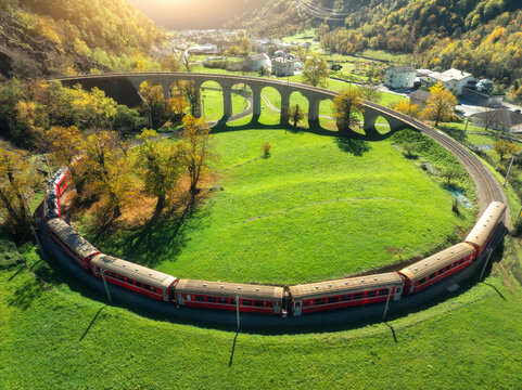 Aerial view of red train on Kreisviadukt in alpine mountains at sunset in autumn. Bernina Express, Switzerland. Top view of train, railroad, green grass , colorful trees in fall. Brusio spiral viaduct