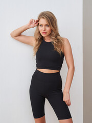 Naklejka premium Young woman in a black athletic outfit poses confidently, hand touching her hair. Her top is sleeveless and paired with bike shorts. Her expression is calm and focused. Casual and stylish