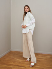 Smiling woman with long brown hair in a white oversized shirt, beige sweater vest, and pleated wide-leg pants stands on a hardwood floor wearing white heeled sandals