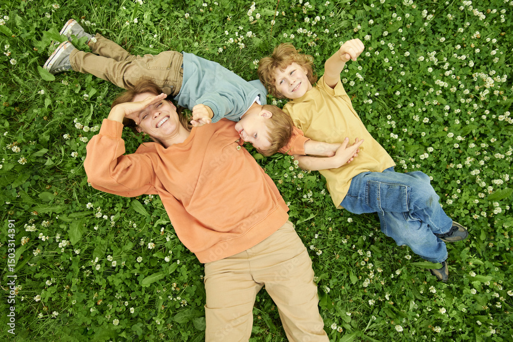 Wall mural candid top view of happy mother lying with two sons on clover field and smiling up at camera playful