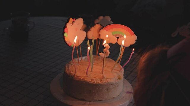 A little child girl in a hat celebrates her third birthday at home with balloons while eating a birthday cake with her relatives.
