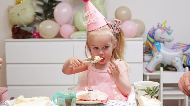 A little child girl in a hat celebrates her third birthday at home with balloons while eating a birthday cake with her relatives.