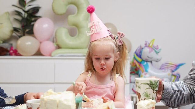 A little child girl in a hat celebrates her third birthday at home with balloons while eating a birthday cake with her relatives.