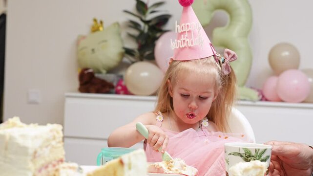 A little child girl in a hat celebrates her third birthday at home with balloons while eating a birthday cake with her relatives.