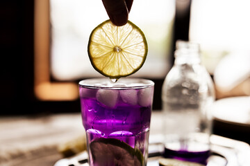 Hand holding a lime slice with tonic dripping into an eye-catching purple cocktail, alongside a...