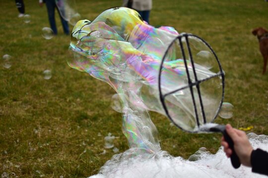 Colorful soap bubbles floating in the air with children playing in the background. Outdoor fun on a grassy field, creating giant bubbles with a wand. Joyful childhood scene in motion.
