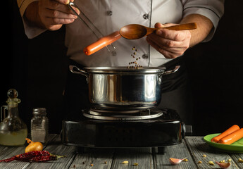 A chef is skillfully handling sausages and pouring spices into a pot on a stovetop. The kitchen is dimly lit, creating a cozy atmosphere while meal preparation takes place