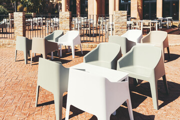 Outdoor seating area with modern plastic armchairs in neutral tones arranged around white tables on a sunny terrace with brick flooring and a stylish stone-and-metal fence in the background