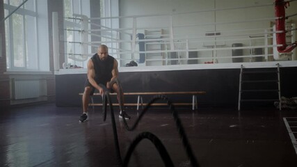 Black sportsman exercises with battle ropes near boxing ring - Powered by Adobe