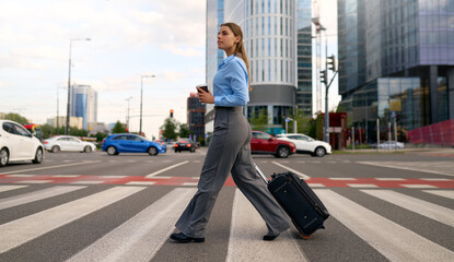 Confident businesswoman walking across city crosswalk with suitcase and coffee