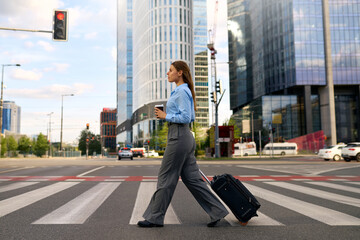 Confident businesswoman walking across city crosswalk with suitcase and coffee