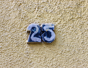 Street address numbers, number 25, painted in white on a raised blue background set on a rough yellow stucco wall.