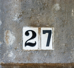 Street address number, number 27, in black on white tile inset into a dark rough stone wall.