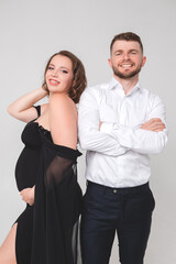 Beautiful pregnant young girl in a black long dress with her husband on a white background in a photo studio. Pregnancy.