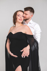 Beautiful pregnant young girl in a black long dress with her husband on a white background in a photo studio. Pregnancy.