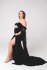 Beautiful pregnant young girl in a black long dress on a white background in a photo studio....