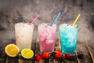 Close-up of glasses with ice surrounded by smoke, filled with colorful soft drinks of various flavors, accompanied by the fruits used in them: lime, lemon, strawberries, raspberries, and blackberries.