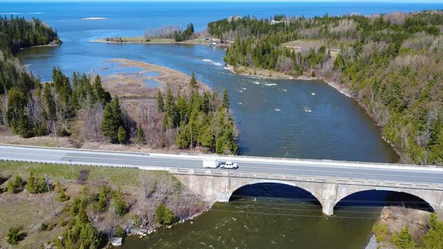 Panoramic aerial view of the mouth of the Mitis River into the St. Lawrence River and its double-arched road bridge crossed by a truck and its trailer. Quebec, Canada, 2025.