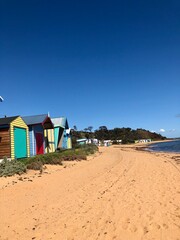 Colorful beach huts line a quiet shoreline under an overcast sky, with waves gently lapping at the sand.