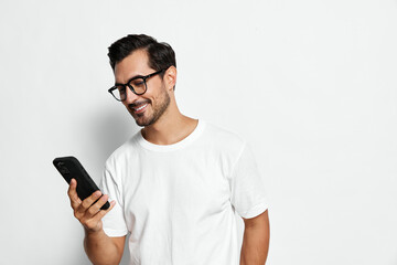 Smiling man wearing glasses and a white t-shirt, looking at his mobile phone with joy, standing...