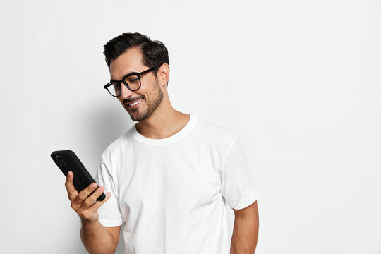 Smiling man wearing glasses and a white t-shirt, looking at his mobile phone with joy, standing against a plain solid background. People lifestyle concept.
