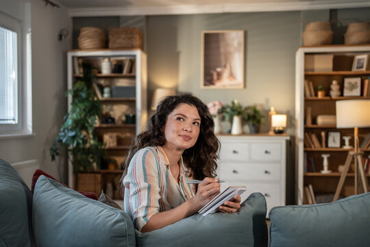 Young woman taking notes on sofa and thinking in cozy living room