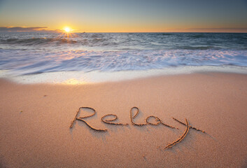 Relax word handwritten on sandy beach at sunset.