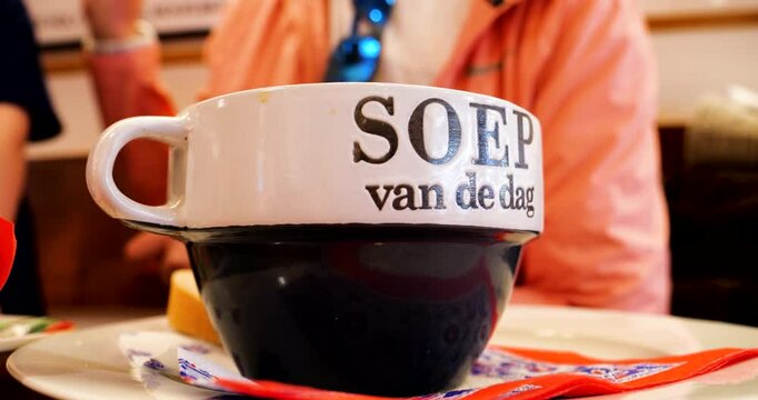 A large ceramic bowl with the words soep van de dag sits on a table in a Texel restaurant, accompanied by bread, butter, and a person wearing orange