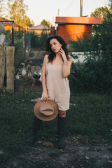 A beautiful woman in a light jumpsuit with a hat stands near a paddock with geese village. tinting