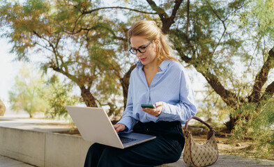 A businesswoman is working outdoors on her laptop while also utilizing her smartphone