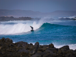 Surfer rides along the face of a curling wave framed by a volcanic coastline and deep blue water. Regular footed rider shows off skills and manoeuvres while surfing waves in the scenic Canary Islands.