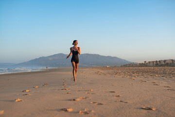 Barefoot woman jogging along wide sandy beach at sunrise, mountain backdrop, long trail of footprints, healthy outdoor workout, serene coastal landscape