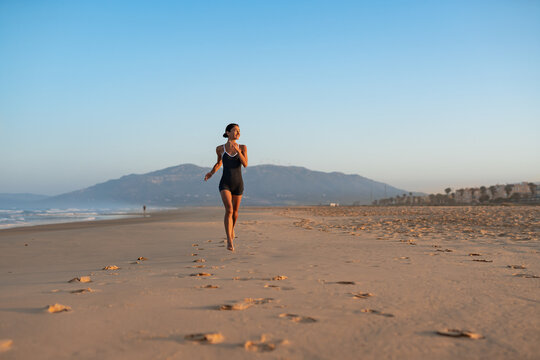 Barefoot woman jogging along wide sandy beach at sunrise, mountain backdrop, long trail of footprints, healthy outdoor workout, serene coastal landscape - Powered by Adobe