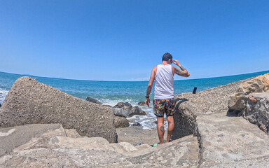 Fototapeta premium Man walks on a cliff overlooking the Mediterranean Sea. The man wears a white tank top and a floral costume. The photo was taken during a sunny day in Sicily