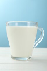 Fresh milk in glass cup on white wooden table against light blue background, closeup