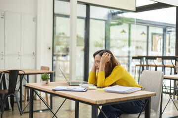 A woman is sitting at a desk with a laptop and a stack of papers. She is looking at the laptop and...