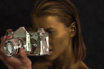 CPortrait of beautiful young woman with face and skin painted gold and holding ceramic silver camera statue for decoration, while pretending taking a picture. Fine art portrait concept