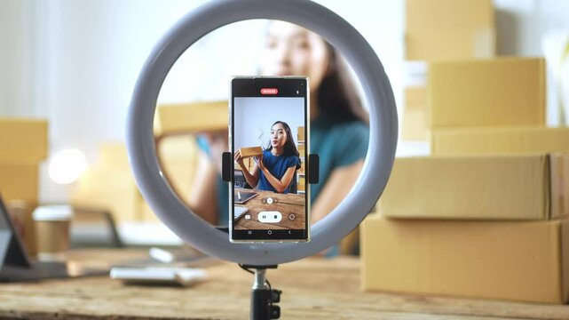 A woman is taking a picture with a phone that is attached to a ring light. The ring light is in front of a stack of cardboard boxes