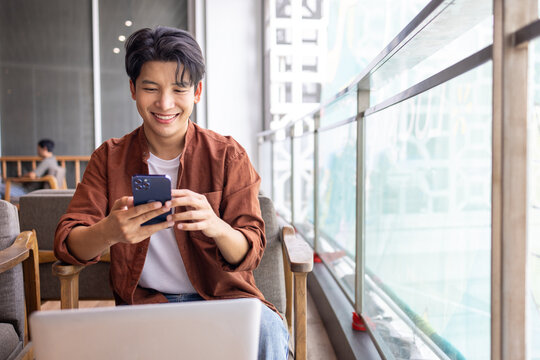 Happy Asian man using mobile phone and working with computer laptop while sitting in coffee shop