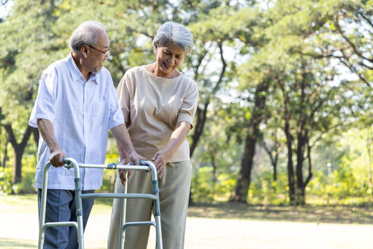 Asian senior husband take care to wife having knee pain while walking with walker stick in the park, Physical therapy in outdoor concept