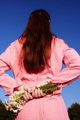A young woman holding flowers behind her back, wearing a pink outfit, with long brown hair, standing against a bright blue sky, evoking feelings of innocence and joy